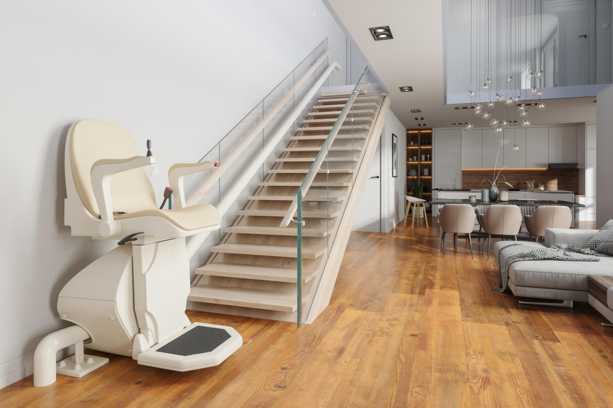 A modern home interior featuring a stair lift installed on a staircase with glass railings, leading to an upper floor. The stair lift has a beige seat and armrests.