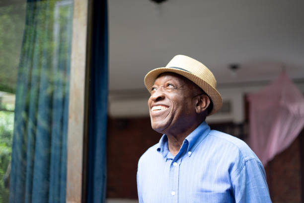 Smiling elderly man wearing a hat and a blue shirt, standing near a window, representing senior citizens and long-term care in New Jersey.