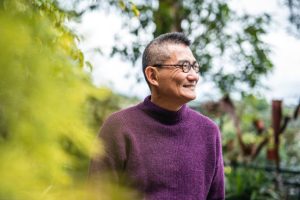 "A smiling senior woman wearing glasses and a purple sweater, standing outdoors in a natural setting."