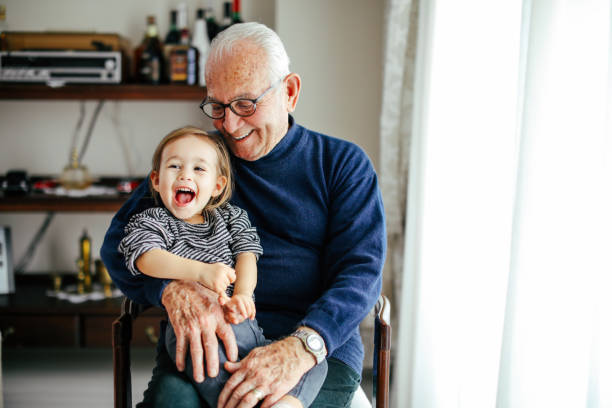 Elderly man wearing glasses, smiling as he holds a joyful young child on his lap in a cozy home setting.

