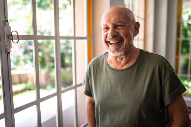 "A joyful senior man wearing a green t-shirt, smiling brightly while standing near a sunlit window."

