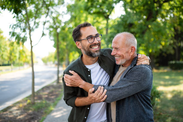 "A smiling younger man with glasses embraces an older man in a warm hug while standing outdoors on a tree-lined pathway, symbolizing care and support."

