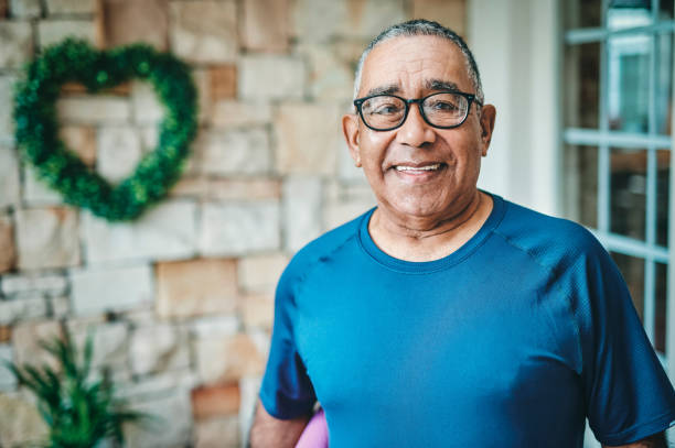 Smiling elderly man wearing glasses and a blue shirt, standing indoors with a heart-shaped plant decoration in the background, representing participants in New Jersey’s Long-Term Care Partnership Program.