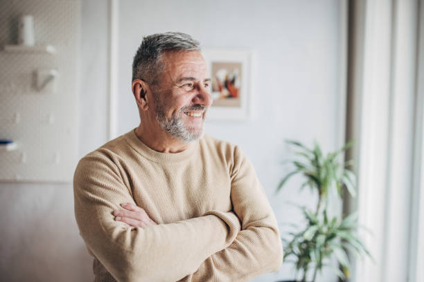 A smiling senior man standing with folded arms in a well-lit home, appearing content and relaxed.