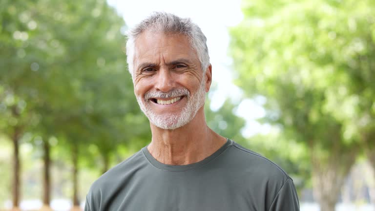 Smiling older man with gray hair standing outdoors in a green park, representing health and vitality.

