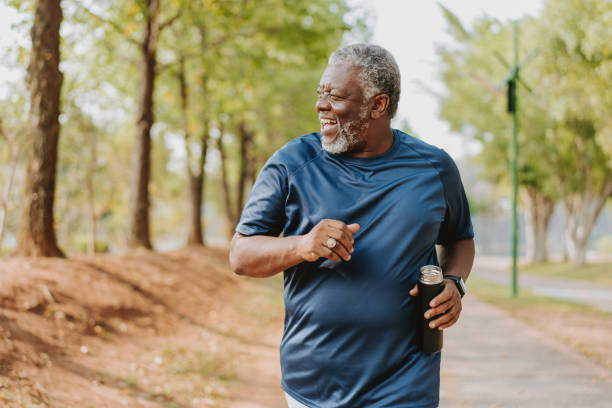 Smiling older man jogging on a tree-lined path, holding a water bottle, symbolizing active living and well-being.

