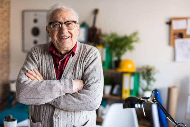 Elderly man wearing glasses and a cozy cardigan, smiling confidently in a home office setting, representing comfort and assurance.

