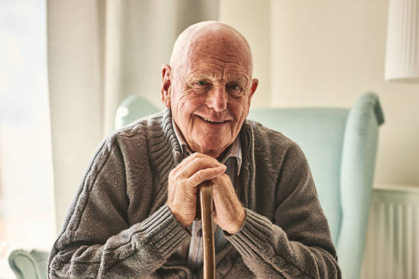 Elderly man smiling while sitting in a cozy living room, leaning on a wooden cane, representing comfort and well-being.

