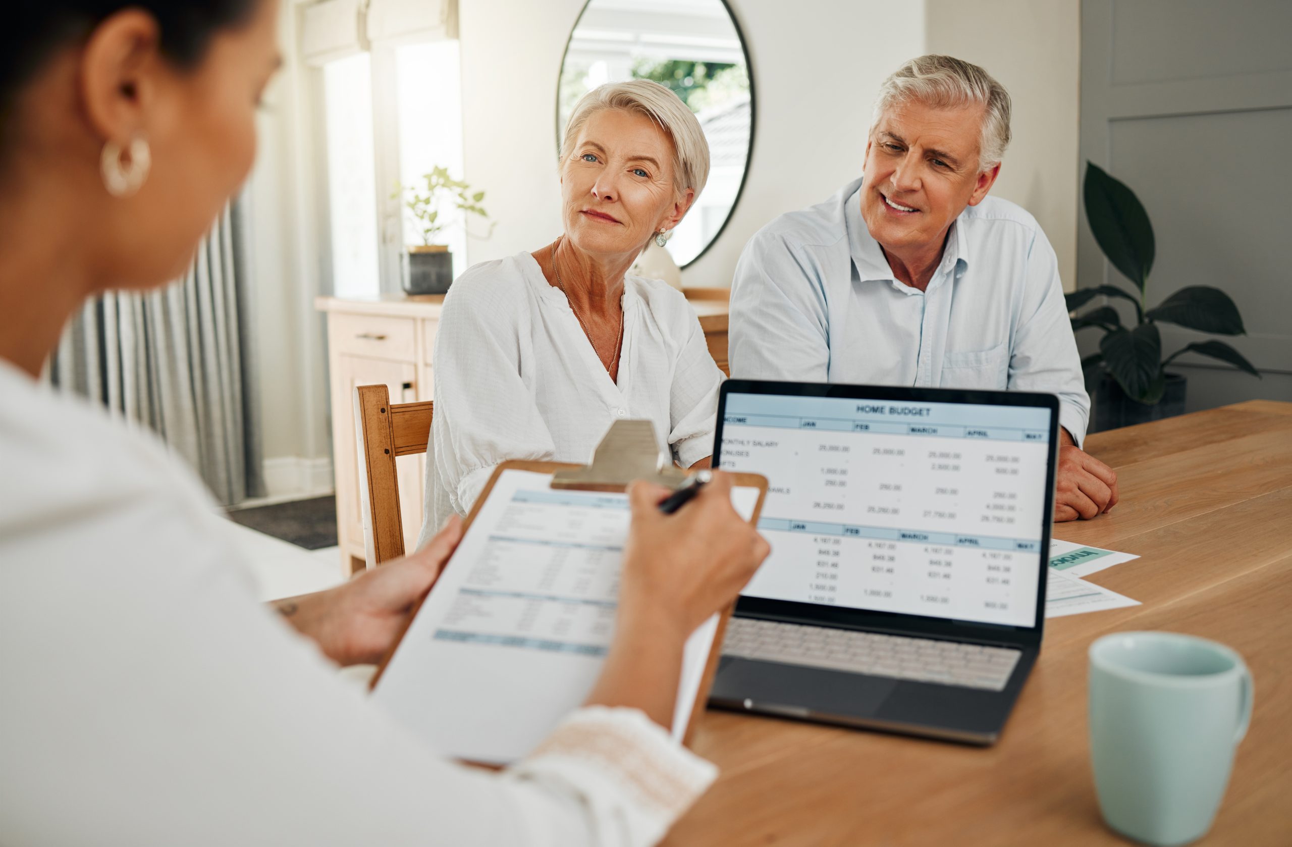 A financial advisor discussing Medicare costs with an elderly couple at home. A laptop screen displaying a budget breakdown is visible in the foreground.