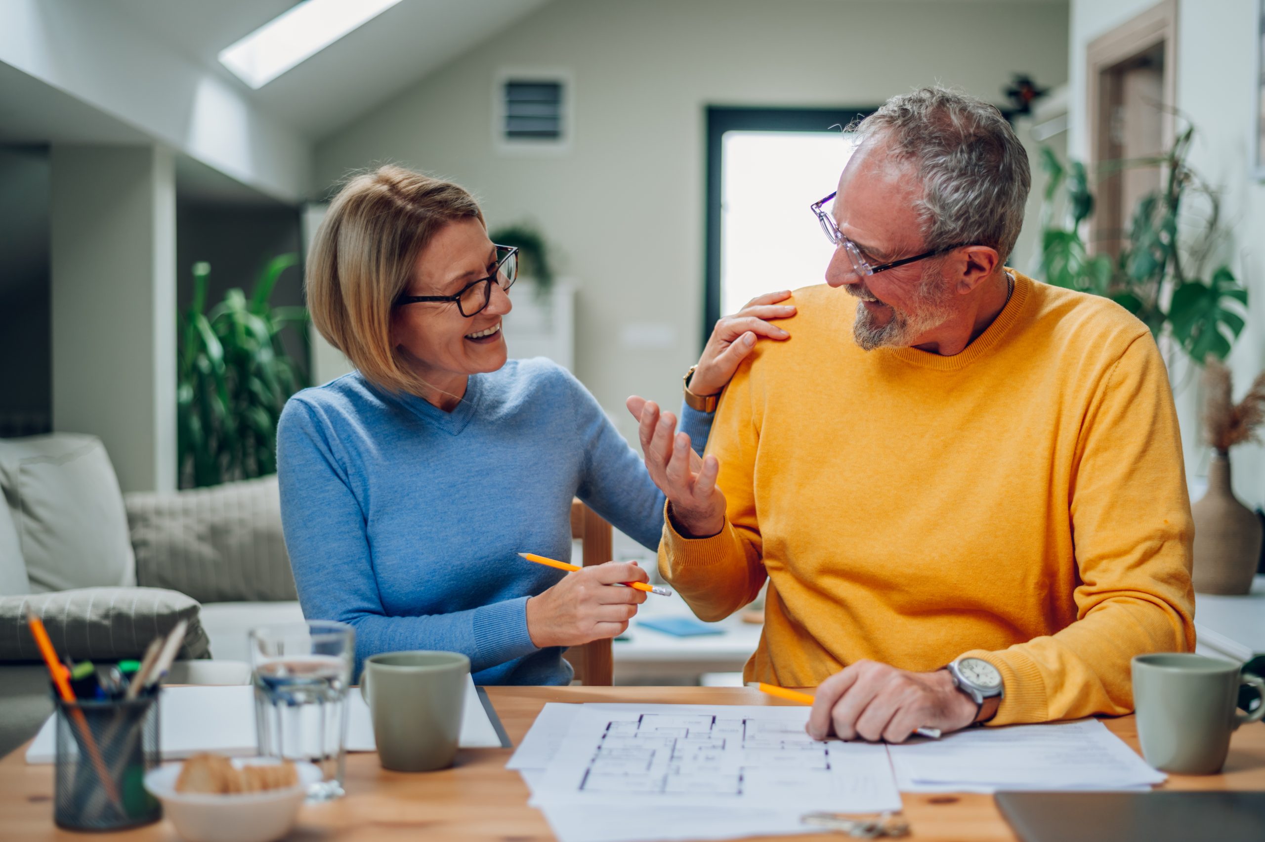 A senior couple sitting at a table, smiling and discussing documents related to insurance coverage in a well-lit home setting.

