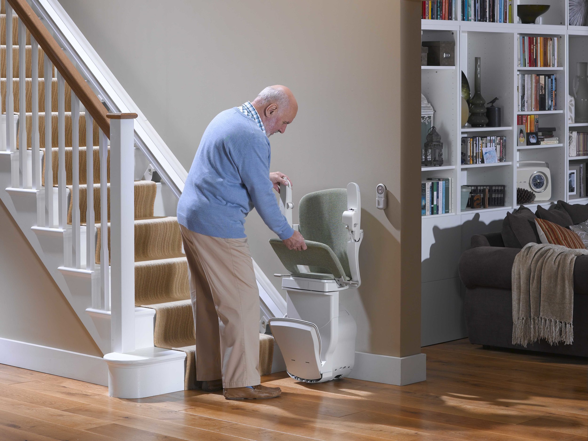 An elderly man in a blue sweater adjusting a stair lift in a cozy living room with a staircase and bookshelves in the background.