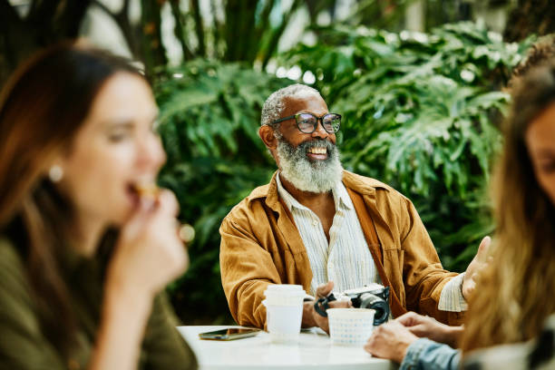 "Smiling elderly man wearing glasses and a brown jacket, sitting at an outdoor café with friends, enjoying a conversation."

